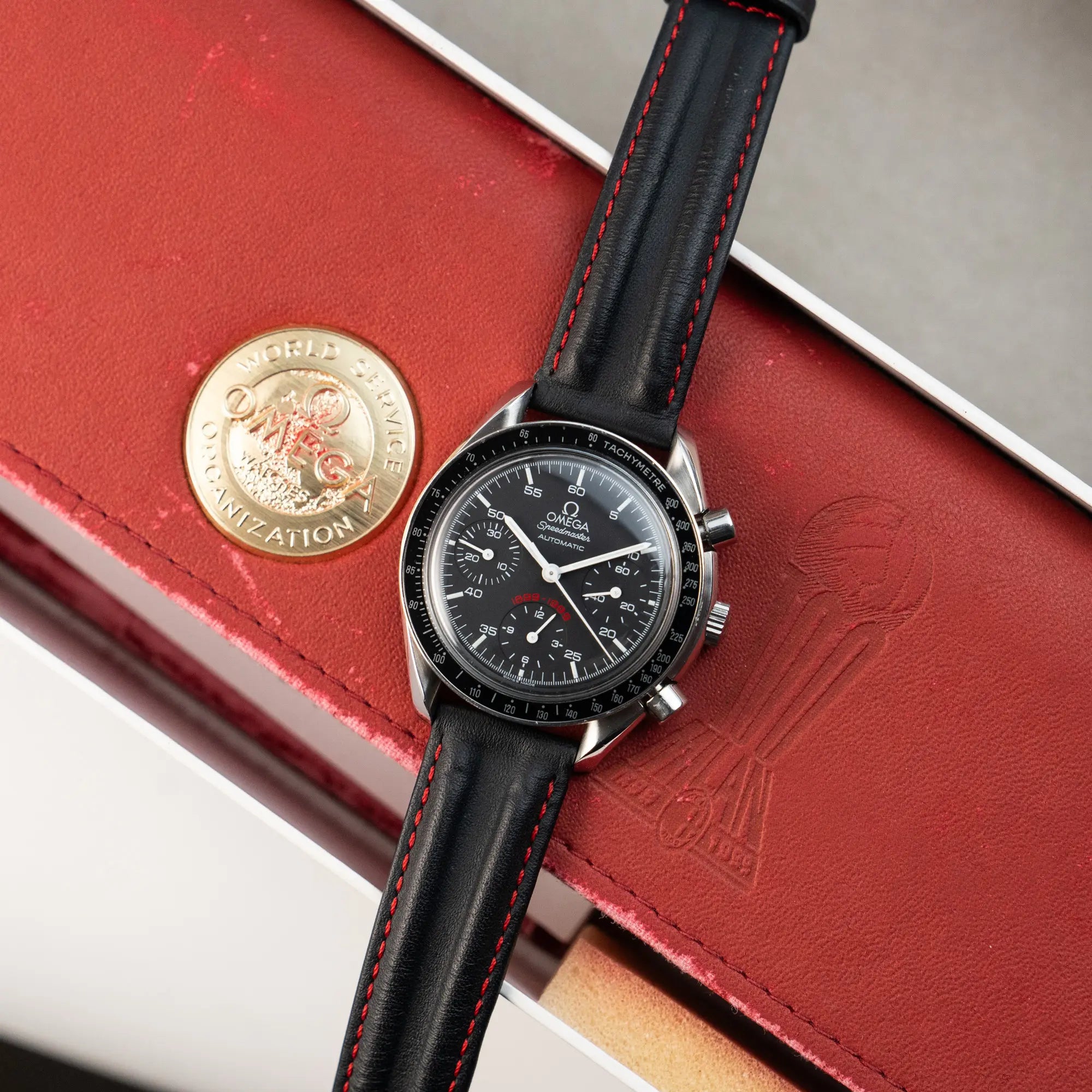 Produktfotografie der Omega Speedmaster Reduced AC Mailand 100th Anniversary Limited Edition zusammen mit der Uhrenbox