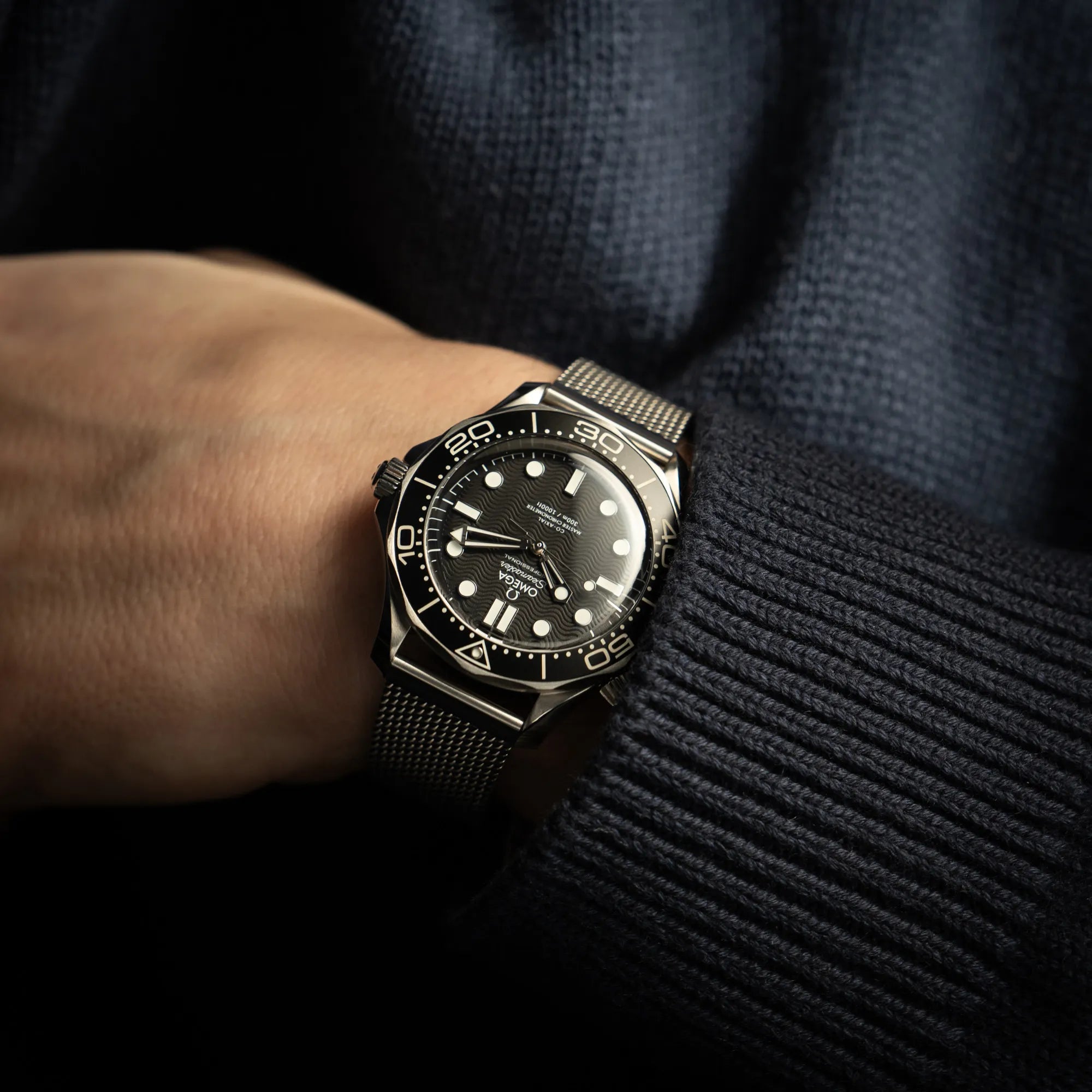 Mann in blauem Strickpullover trägt die Omega Seamaster 300M Diver 210.30.42.20.01.010 mit dems schwarzen Zifferblatt im Wellen-Design am Milanese-Edelstahl-Armband an seinem Handgelenk