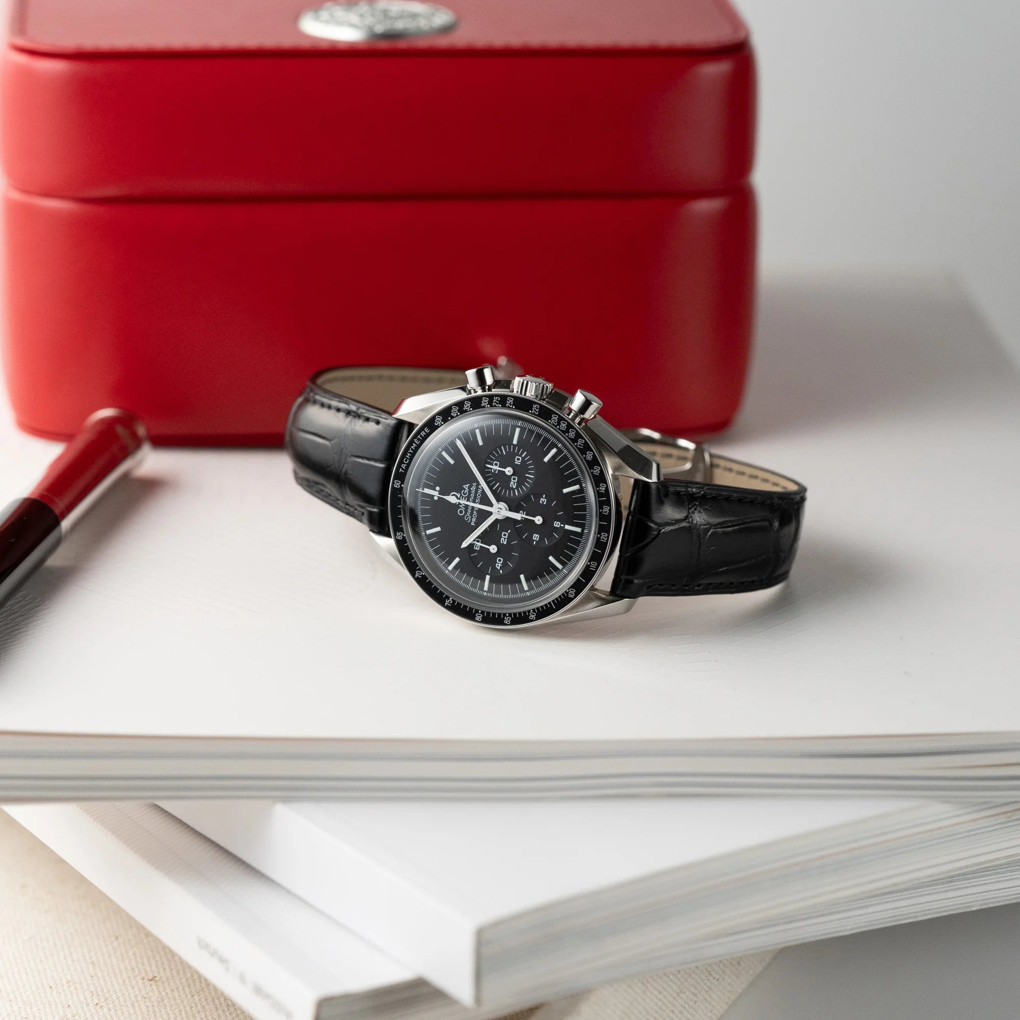 Produktfotografie der Omega Speedmaster Moonwatch Professional am schwarzen Lederband vor der roten Omega-Uhrenbox