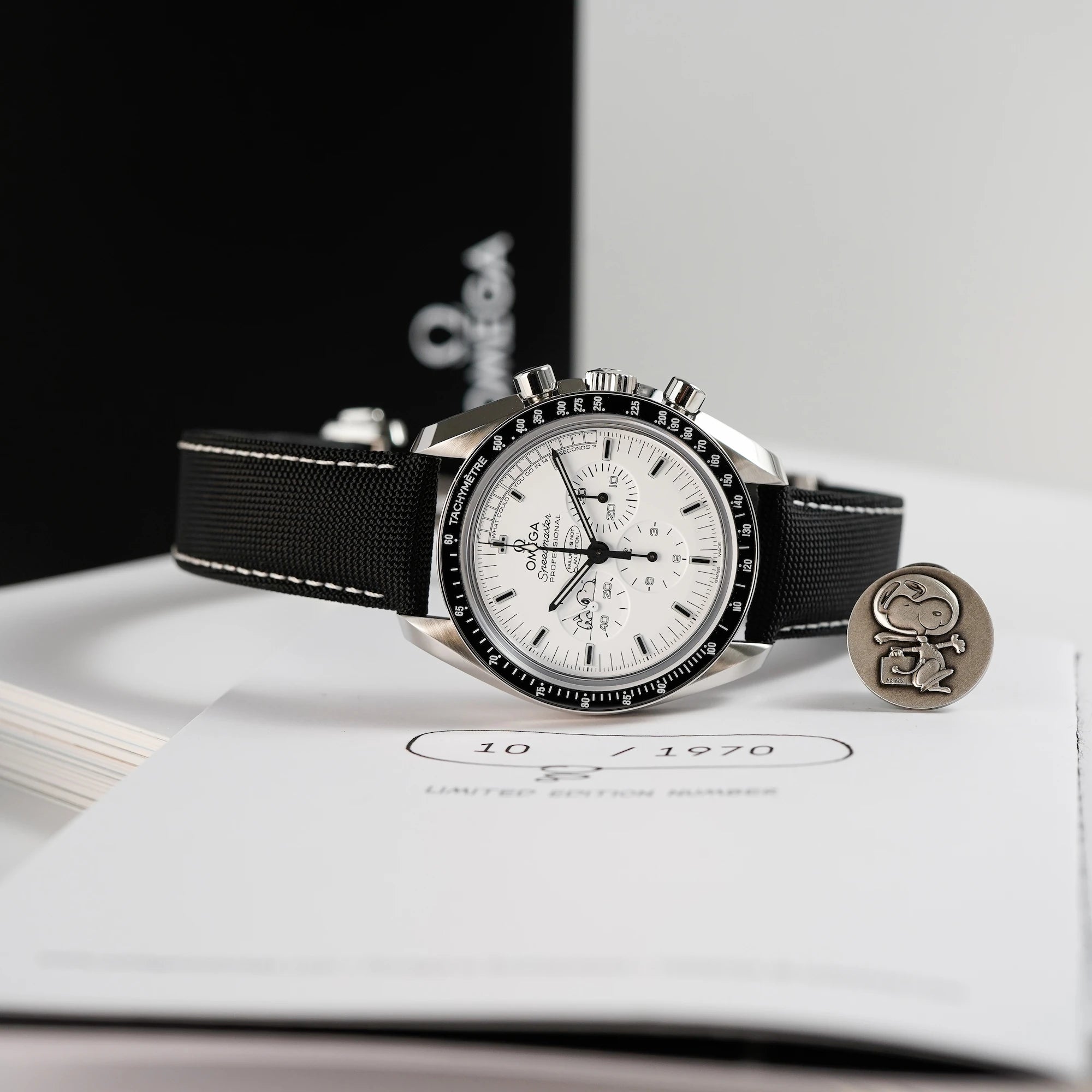 Produktfotografie der limitierten Omega Speedmaster Moonwatch Silver Snoopy mit dem weißen Zifferblatt zusammen mit dem Snoopy-Pin aus dem Lieferumfang