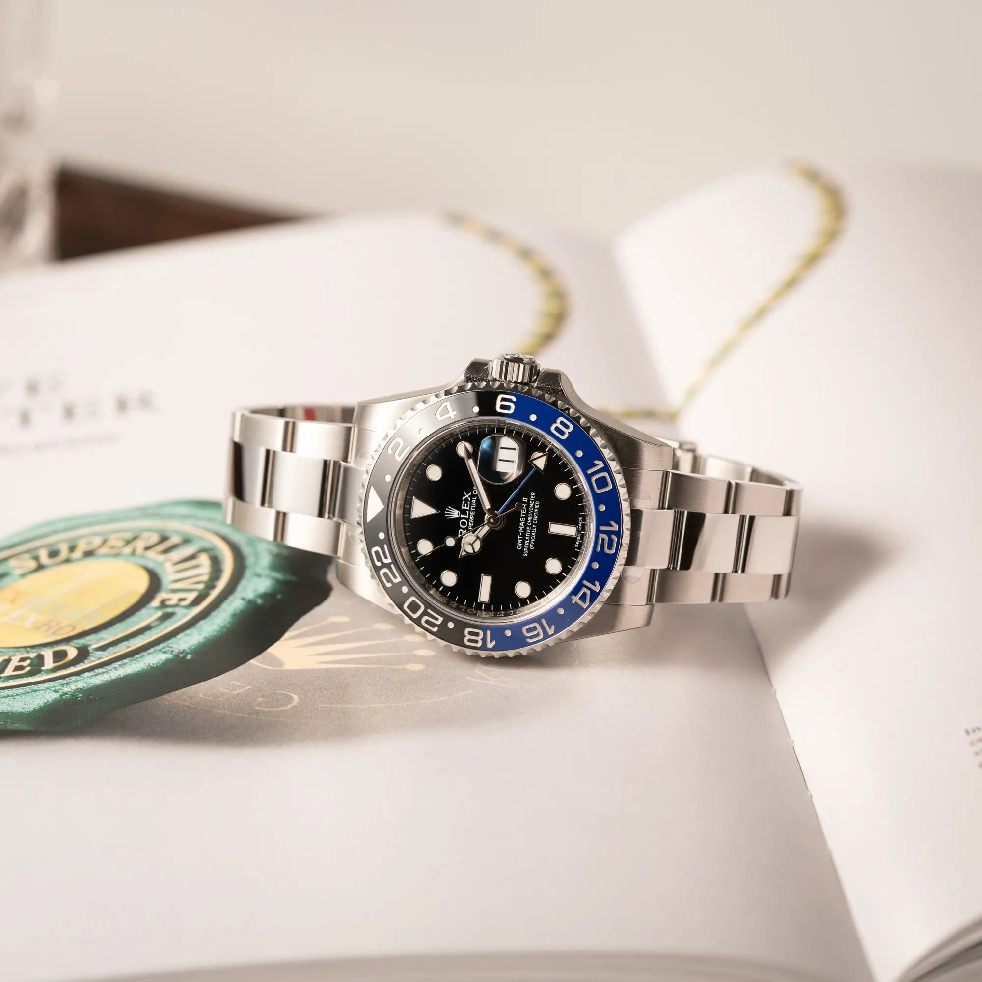 Rolex GMT-Master II der Referenz 116710BLNR mit dem Nickname "Batman" mit der schwarz blauen Keramik-Lünette am Oyster-Stahlband im verklebten Neuzustand liegt auf einem Buch, das ein Rolex Hangtag zeigt