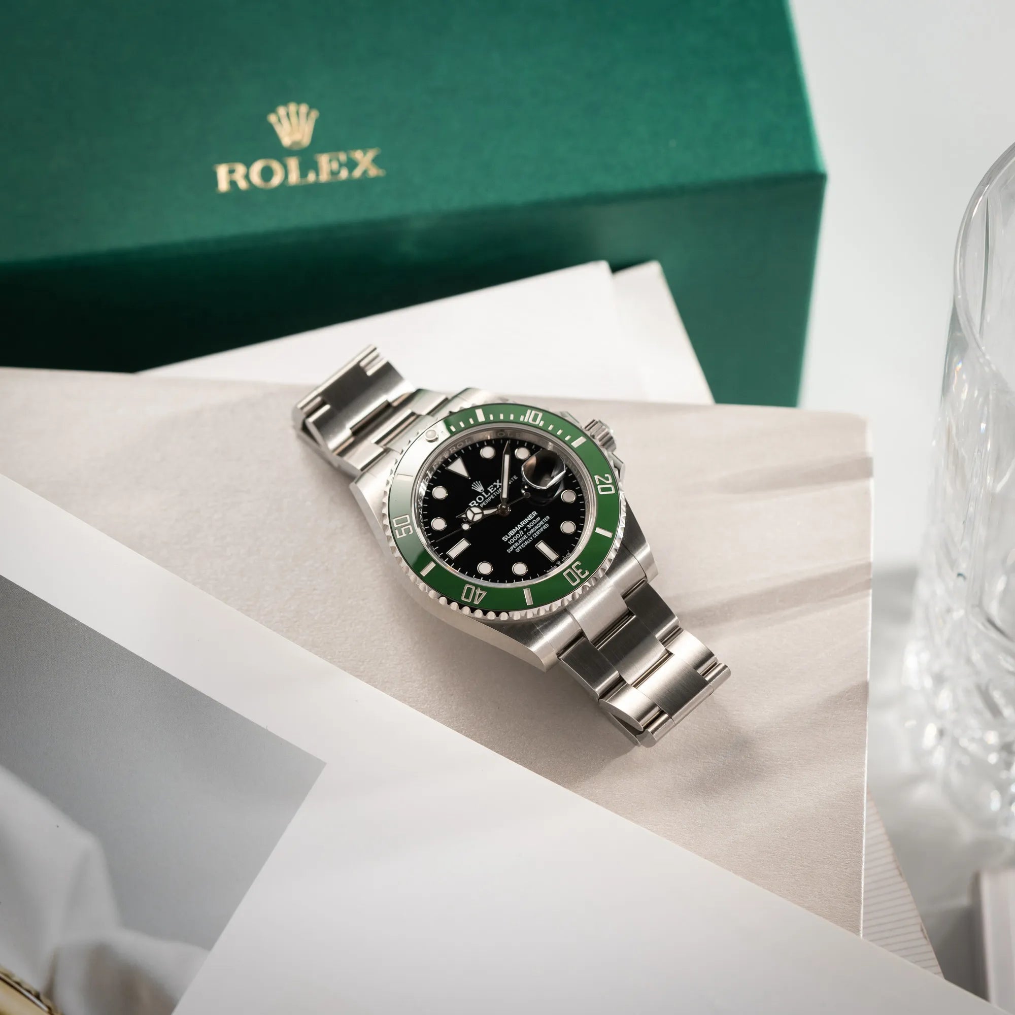 Produktfotografie der Rolex Submariner Date Starbucks 126610LV Mark 2 aus 2025 mit der typischen grünen Cerachrom-Lünette neben dem in 2025 neuen grünem Umkarton