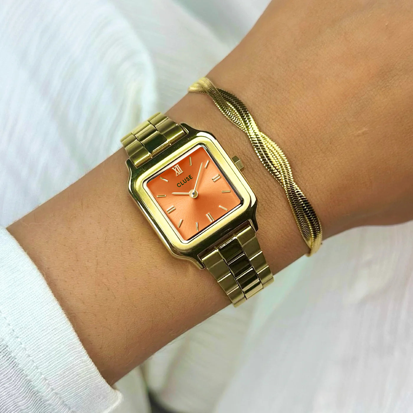 Frau trägt die Cluse Stahl-Uhr "Gracieuse Petite" mit einem gelbgold-farbenem Gehäuse und orangem Zifferblatt an ihrem Handgelenk und kombiniert diese mit einem goldenen Armband