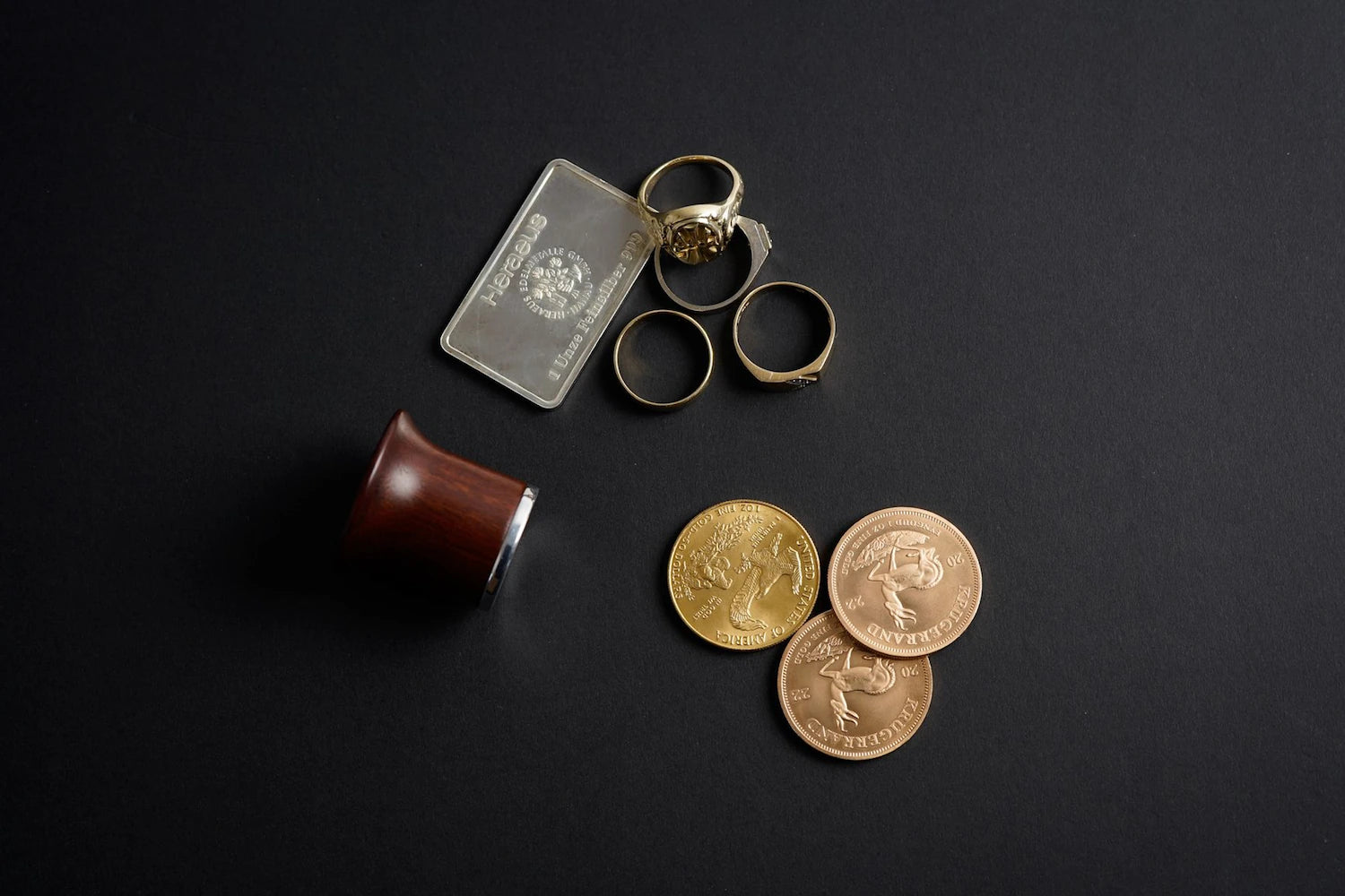 Silberbarren, Goldmünzen und alte Ringe liegen während eines Goldankaufs zusammen mit einer Lupe auf schwarzem Hintergrund