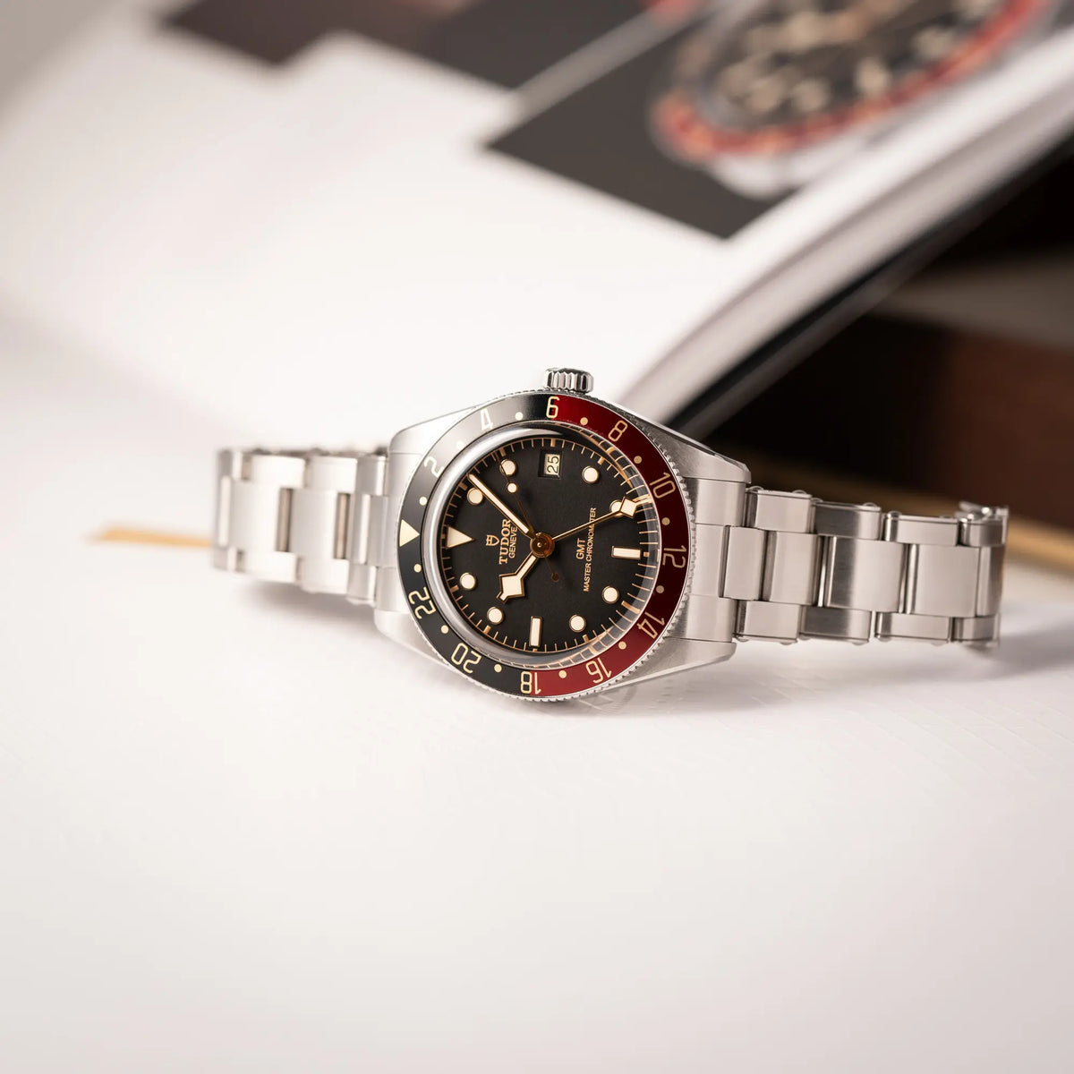 Tudor BlackBay GMT Coke der Referenz 7939G1A0NRU mit der schwarz-roten Lünette und bronzefarbenen Details am Edelstahlarmband  liegt seitlich auf dem Uhrengehäuse auf einem Uhrenbuch