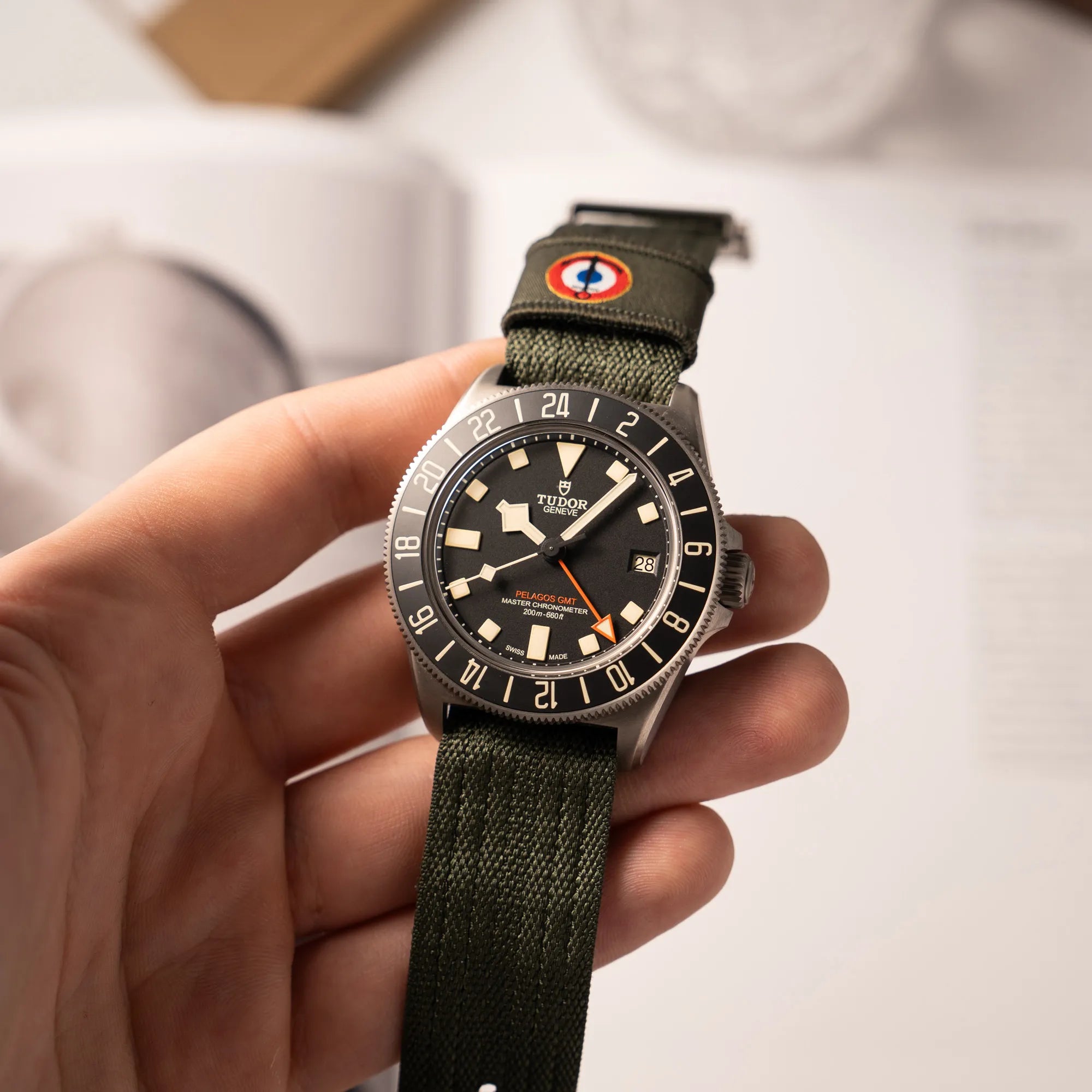 Mann hält die Tudor Pelagos GMT Military der Referenz M2542G257NU-0002 mit Titan-Gehäuse und schwarzem Zifferblatt an einem olivgrünen Nato-Strap in seiner Hand und präsentiert die Armbanduhr in die Kamera