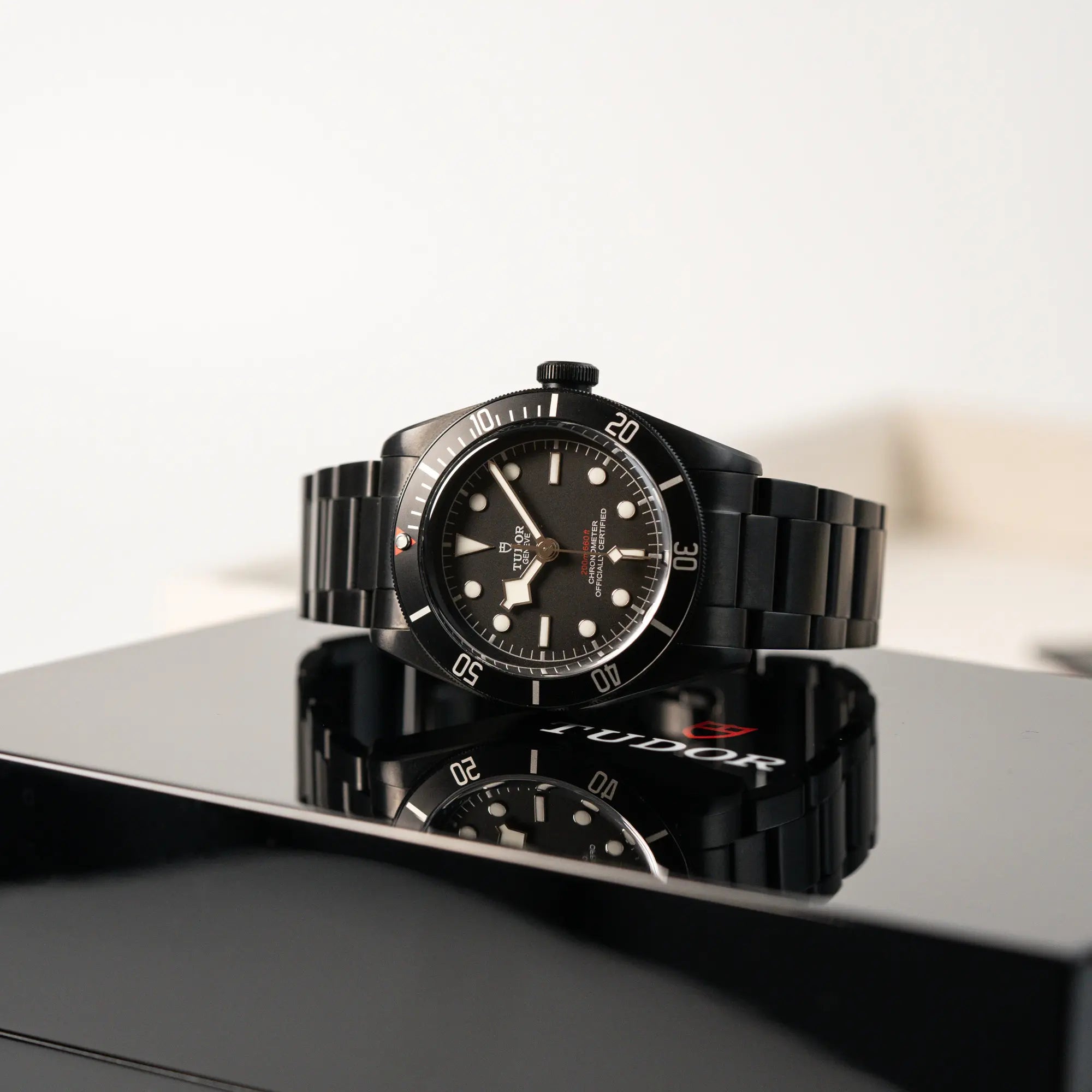 Produktfotografie der Tudor Black Bay Dark 79230DK mit einem schwarz beschichteten Gehäuse und weißen Ziffern & Indizes, während deine Uhr auf der schwarz glänzenden Uhrenbox liegt