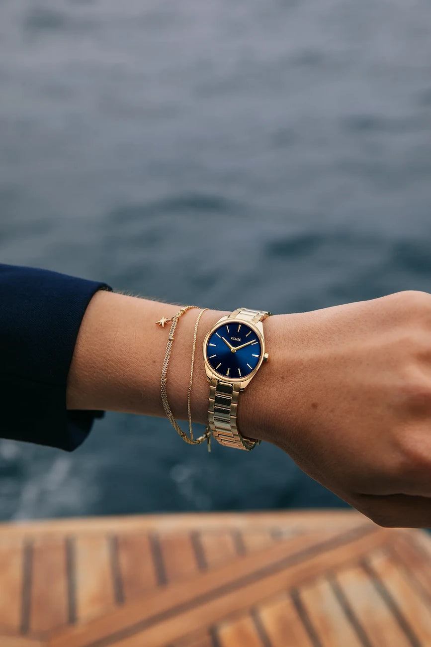 Wristhot der Feroce Uhr von Cluse mit goldfarbenem Gehäuse mit blauem Zifferblatt auf einem Steg vor einem See