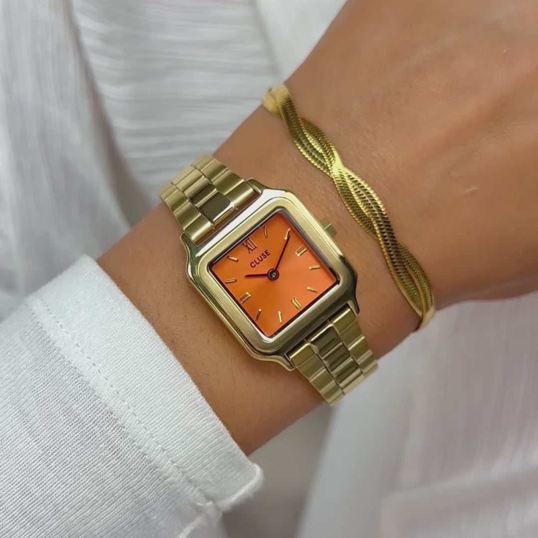 Frau präsentiert die Cluse Stahl-Uhr "Gracieuse Petite" mit einem gelbgold-farbenem Gehäuse und orangem Zifferblatt mit einem Wrisrtoll